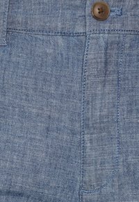 Tessuto di denim azzurro chiaro con una sottile texture, caratterizzato da un bottone marrone e dettagli di cucitura. Include una tasca frontale e linee di cucitura diritte.