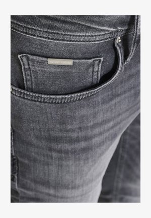 Primer plano del bolsillo de unos vaqueros de mezclilla gris oscuro con una pequeña etiqueta metálica rectangular pegada cerca del borde del bolsillo.