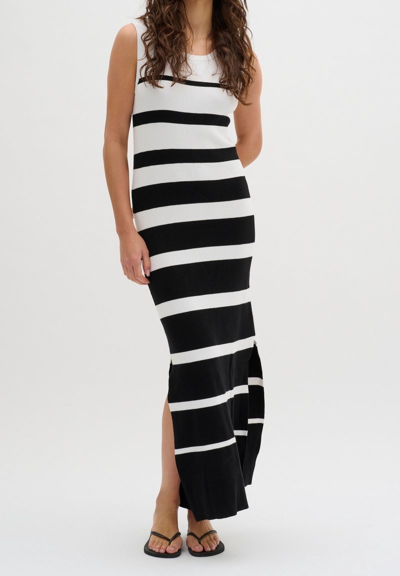 Femme portant une robe sans manches, rayée noire et blanche, longueur cheville, avec des fentes latérales, debout avec une main derrière le dos.