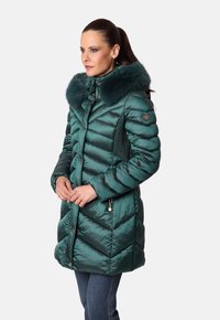 Mujer con un abrigo de invierno acolchado en color verde azulado y capucha con borde de piel, de pie con las manos entrelazadas, mirando hacia la izquierda sobre un fondo liso.