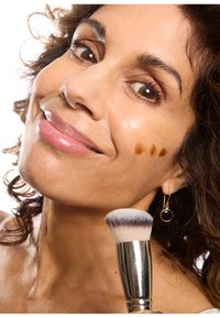 Pennello per il trucco con setole bianche e nere tenuto da una persona mentre applica un liquido marrone sulla guancia. Orecchino a cerchio dorato visibile. Capelli ricci.