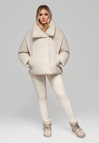 Béžová péřová bunda s velkým límcem a předními kapsami, v kombinaci se smetanovými legínami a béžovými šněrovacími kotníkovými botami na neutrálním pozadí.