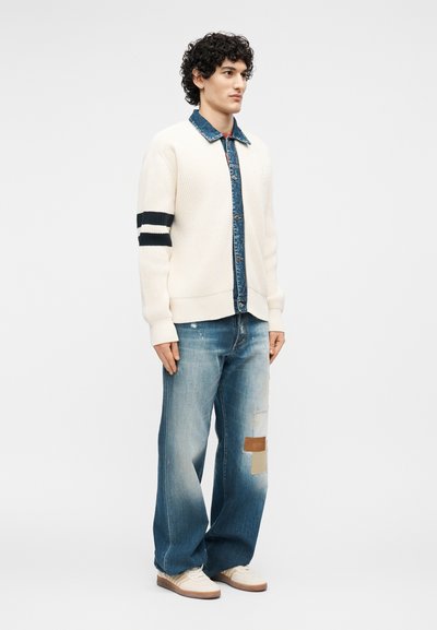Jeune homme portant un cardigan en maille crème à rayures noires, une chemise en denim, un jean large patchwork et des baskets blanches sur fond blanc.
