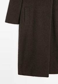 Detail rukávu a boční kapsy tmavohnědého vlněného kabátu na bílém pozadí, ukazující strukturu a rovný lem.