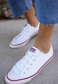 Białe kanwasowe trampki z czerwonymi akcentami, gumową osłoną palców i sznurowaniem. Zawierają metkę Converse All-Star na języku.