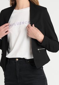 Musta bleiseri, jossa on istuva leikkaus ja vetoketjuyksityiskohtia, päällä valkoinen t-paita, jossa on vaaleanpunaisella kirjoitettu sana "INFLUENCE".