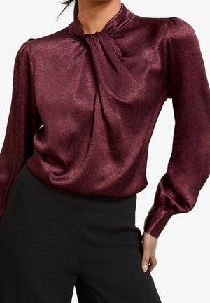 Vrouw draagt een glanzende bordeauxrode blouse met lange mouwen en een geplooide halslijn, gecombineerd met zwarte hoge taillebroek, hand rustend op haar heup.