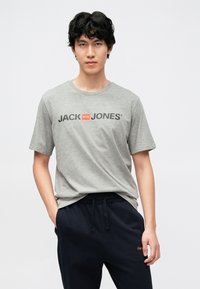Grå T-skjorte med "JACK & JONES" trykket i svart og rødt. Kortermet, rund hals. Kombinert med mørkeblå bukser med snøring i midjen.