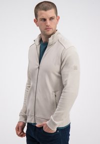 Jasno beżowa kurtka na zamek z teksturowanymi wstawkami na rękawach, dwoma bocznymi kieszeniami oraz stójką, noszona na ciemnoniebieskiej koszuli.