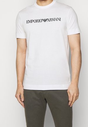 Homme portant un t-shirt blanc à manches courtes avec le texte noir "EMPORIO ARMANI" et le logo, associé à un pantalon vert olive foncé.