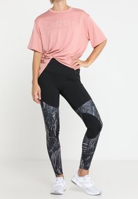 Persona che indossa una maglietta rosa "Just Do It" annodata in vita, leggings neri con fantasia e scarpe da ginnastica bianche, in piedi con una mano su un fianco.
