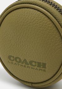 Custodia rotonda in pelle verde oliva con superficie testurizzata e zipper visibile, caratterizzata dalla stampa in rilievo del marchio "COACH LEATHERWARE" su un lato.