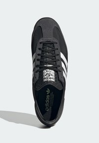 Svart sneakers med texturerad svart överdel, tre vita ränder och en glansig svart tunga med Adidas-logotyp. Snörning.