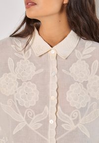 Blusa bianca ricamata con motivi floreali, colletto scallop e chiusura a bottoni, realizzata in un tessuto leggero e trasparente.