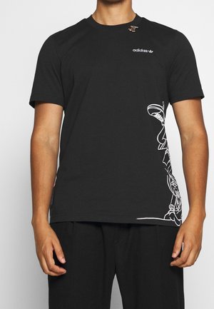 Person iført en sort Adidas T-shirt med hvide grafiske linjer på nederste højre side og en lille nål nær kraven.