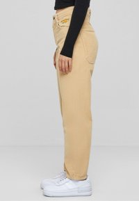 Pantaloni in corduroy beige con design a gamba dritta, dotati di un logo giallo sulla vita, abbinati a sneakers bianche.
