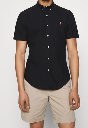 Homme portant une chemise noire à manches courtes avec un petit logo, associée à un short beige jusqu'aux genoux, sur un fond uni.