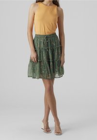En orange ärmlös topp kombinerad med en grön lager-på-lager kjol med blommönster. Kjolen har en dragsko i midjan och en genomskinlig textur.
