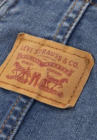 Δερμάτινο εμβλημά της Levi Strauss & Co. με κόκκινη εκτύπωση, που απεικονί ζει άλογα και παντελόνια, ραμμένο σε μπλε τζιν ύφασμα.