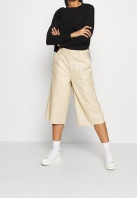 Beige Cropped-Hosen mit strukturiertem Schnitt, aus glattem Stoff gefertigt, mit Vorderfalten und Seitentaschen, kombiniert mit weißen Sneakers.