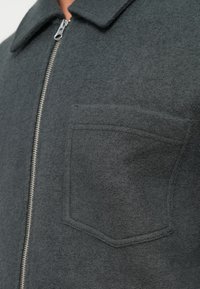 Grå, filtjakke med krage, glidelås og brystlomme. Har teksturert stoff med en subtil overflate som fremhever nytteverdi.