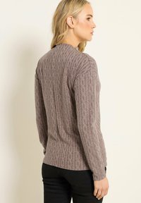 Donna con capelli biondi in profilo laterale che indossa un maglione lungo a maniche con un motivo taupe e pantaloni neri, su uno sfondo chiaro uniforme.