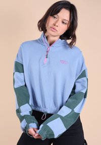 Pull en polaire bleu clair avec fermeture éclair mi-zip, manches à motif vert, poignets élastiques et cordon de serrage à l'ourlet. Comprend un détail de logo.
