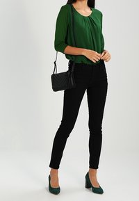 Una donna sta in piedi con sicurezza, indossando una blusa verde, jeans neri e tacchi verdi, mentre tiene una piccola borsa nera contro uno sfondo neutro.