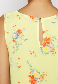 Top sin mangas con estampado floral en tela de color amarillo claro que presenta un cierre en la parte posterior con orificio y una textura suave con colores vibrantes.