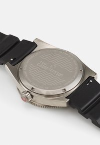 Fond de montre en acier inoxydable avec détails gravés, bracelet en caoutchouc noir, accent rouge sur la couronne, texture lisse, indice de résistance à l'eau indiqué.