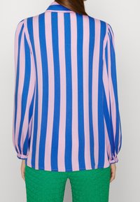 Chemise à manches longues avec col, présentant des rayures verticales rose et bleu dans un tissu texturé. La coupe est décontractée, avec des manches froncées.