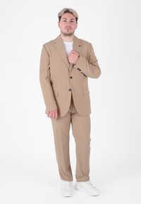 Completo beige con giacca e pantaloni abbinati. Tessuto liscio con una vestibilità su misura. Indossato con una t-shirt bianca e sneakers bianche.