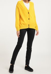 Žlutý kardigan s výstřihem do V, dlouhými rukávy a černými knoflíky. Kombinován s černými slim-fit džíny a černými teniskami. Hladká textura.