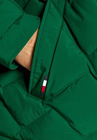 Giacca imbottita verde con motivo a rombi, dotata di una tasca con zip e un'etichetta con logo con accenti rosso, bianco e blu.