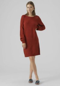 Abito rosso lavorato a maglia con maniche lunghe, scollo a barchetta e una vestibilità comoda, caratterizzato da polsini a costine. Indossato con sandali blu decorati con fiori.
