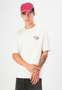 Hvid bomulds T-shirt med korte ærmer med rødt broderet logo. Sorte denim bukser. Rød og sort kasket med synlig syning.