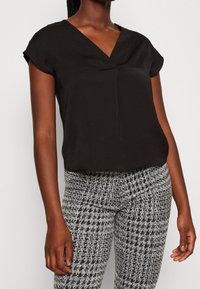 Top corto a maniche corte nero con scollo a V e dettagli di pieghe sottili, abbinato a leggings grigi texturizzati con un motivo a scacchi.