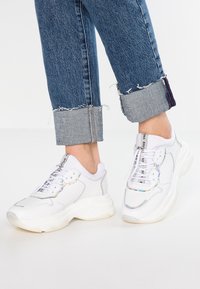 Zapatillas blancas con suela gruesa, que presentan un upper de cuero liso, acentos iridiscentes y cordones blancos. Combinadas con jeans de denim deshilachados.