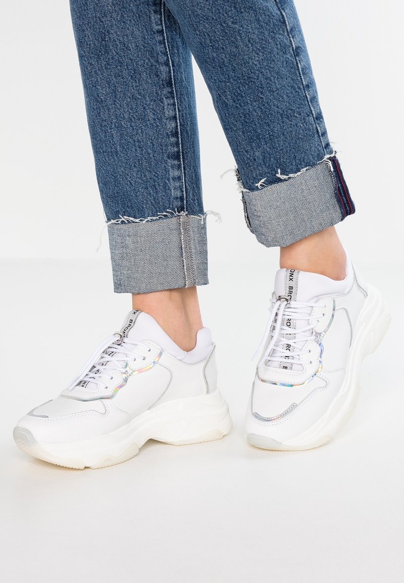 Zapatillas blancas con suela gruesa, que presentan un upper de cuero liso, acentos iridiscentes y cordones blancos. Combinadas con jeans de denim deshilachados.
