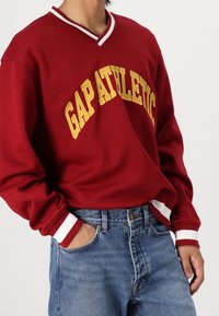 Sudadera roja con el texto amarillo "GAP ATHLETIC", diseño de escote en V y puños acanalados blancos, combinada con jeans de denim azul.