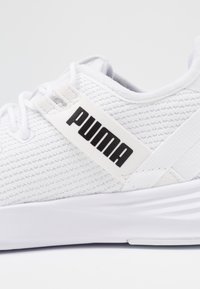 Bílé tenisky Puma s strukturovaným svrškem, plochými tkaničkami a černým logem na pásku. Podešev je solidně bílá a plochá s mírnými drážkami.