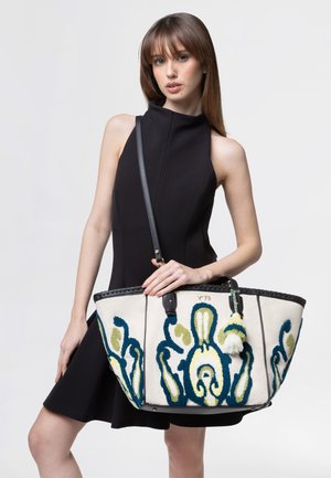 Giovane donna in abito nero senza maniche che tiene una grande borsa bianca con ricamo astratto blu e verde e nappona.