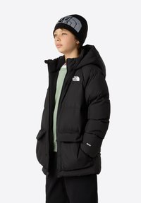 Chaqueta acolchada negra con capucha, que presenta un cierre frontal, dos bolsillos delanteros y detalles de logo; llevada sobre una camisa verde claro y pantalones negros.