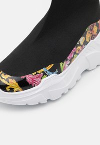 Detailní záběr na černou ponožkovou tenisku s bílou podrážkou a barevným květinovým vzorem na okraji blízko podrážky.