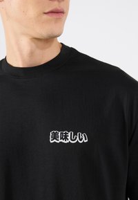 Black cotton t-shirt featuring a white, bold graphic text in Japanese. Round neckline and short sleeves. Smooth texture.