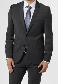 Uomo che indossa un completo grigio scuro con una camicia bianca e una cravatta blu a trama, mani rilassate lungo i fianchi, contro uno sfondo chiaro e uniforme.