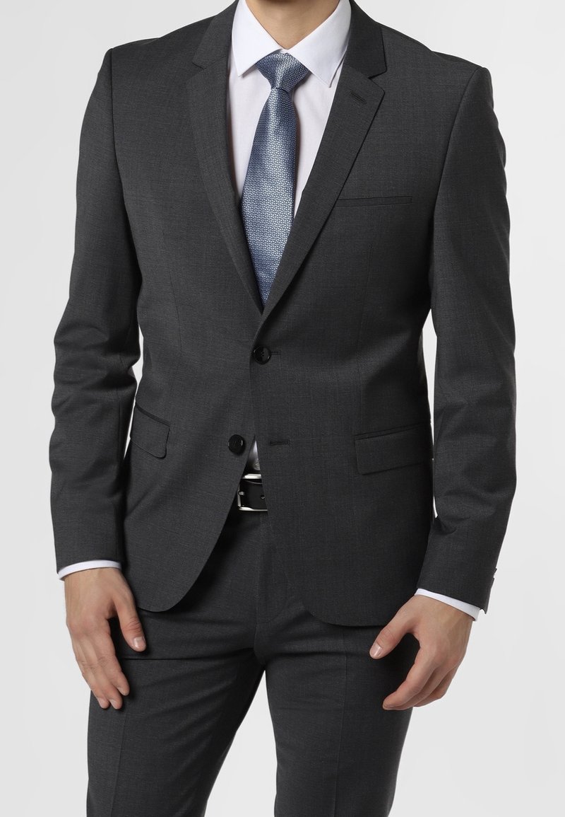 Uomo che indossa un completo grigio scuro con una camicia bianca e una cravatta blu a trama, mani rilassate lungo i fianchi, contro uno sfondo chiaro e uniforme.