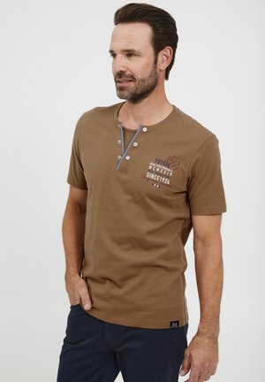 Camisa de algodón marrón de manga corta con cuello redondo, que cuenta con una tapeta de tres botones y detalles gráficos en el pecho. Acentuada con costuras blancas.