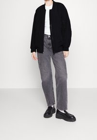 Veste bomber noire à poignets côtelés, portée sur un haut blanc, associée à un pantalon en denim gris taille haute et des mocassins noirs chunky.