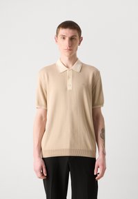Μπεζ πόλο μπλούζα με αντίθετο ανοιχτόχρωμο γιακά, κοντά μανίκια και στενή εφαρμογή. Διαθέτει διακριτική κεντημένη λογότυπο και ριγωτό στρίφωμα.